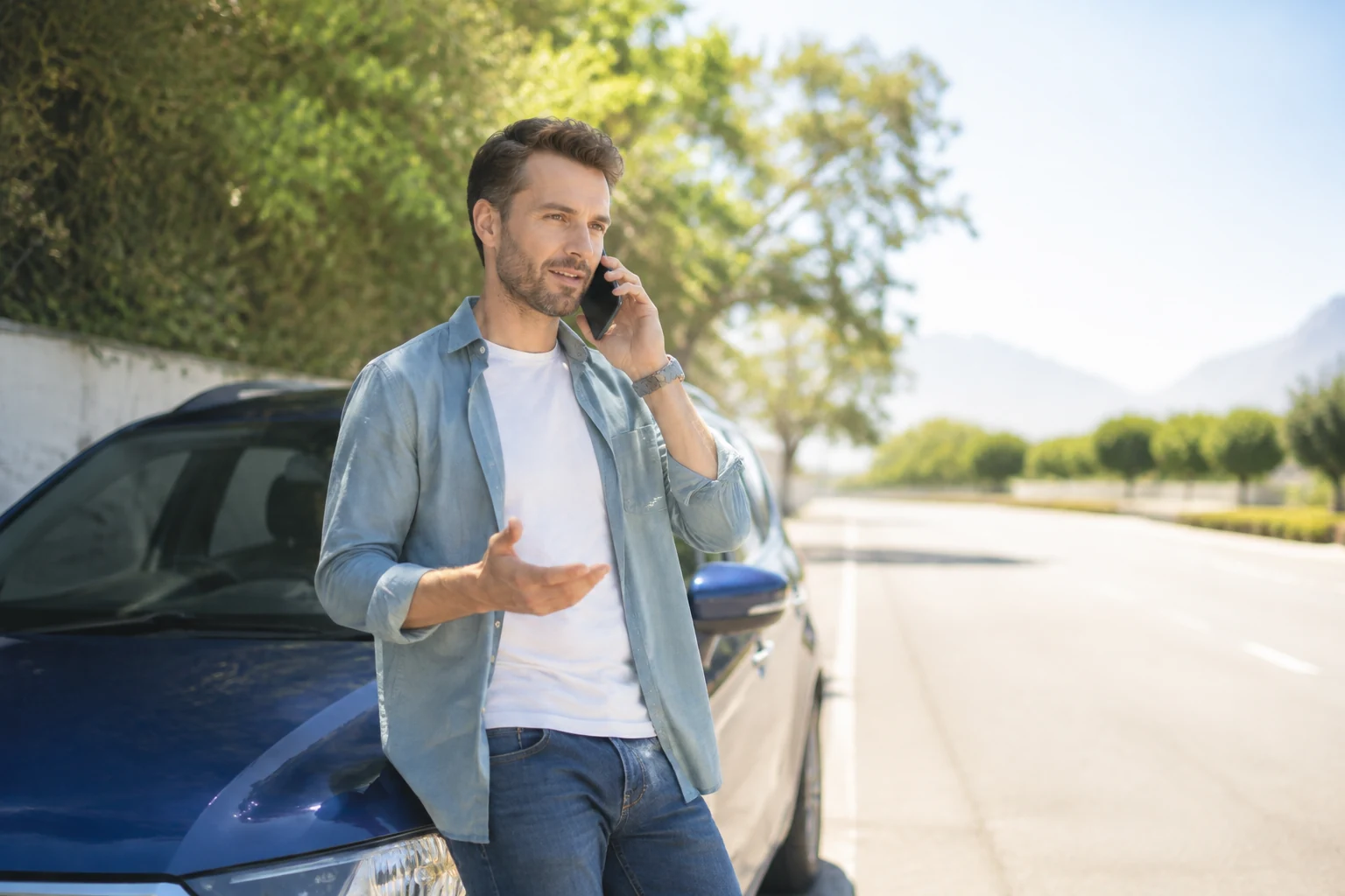 Driver speaking calmly with a support representative on smartphone beside a safely parked car in a bright roadside setting